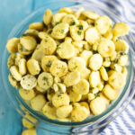 A glass bowl filled with Ranch Oyster Crackers on a striped linen napkin and a bright blue wood background