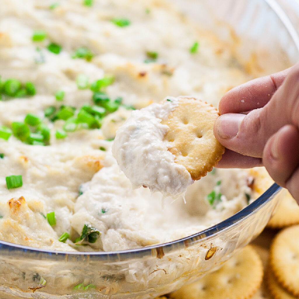 Butter cracker being dipped into a large glass bowl full of crab rangoon dip topped with sliced green onions