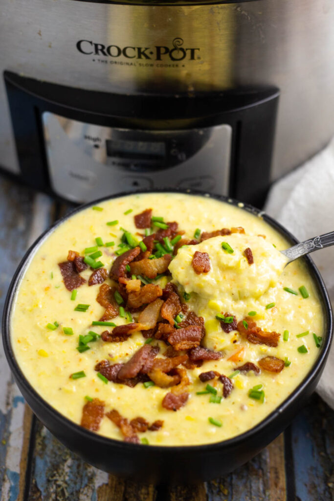 Black ceramic bowl in front of a slow cooker filled with Crock Pot Corn Chowder with a spoon in it, topped with crumbled bacon and chopped chives.