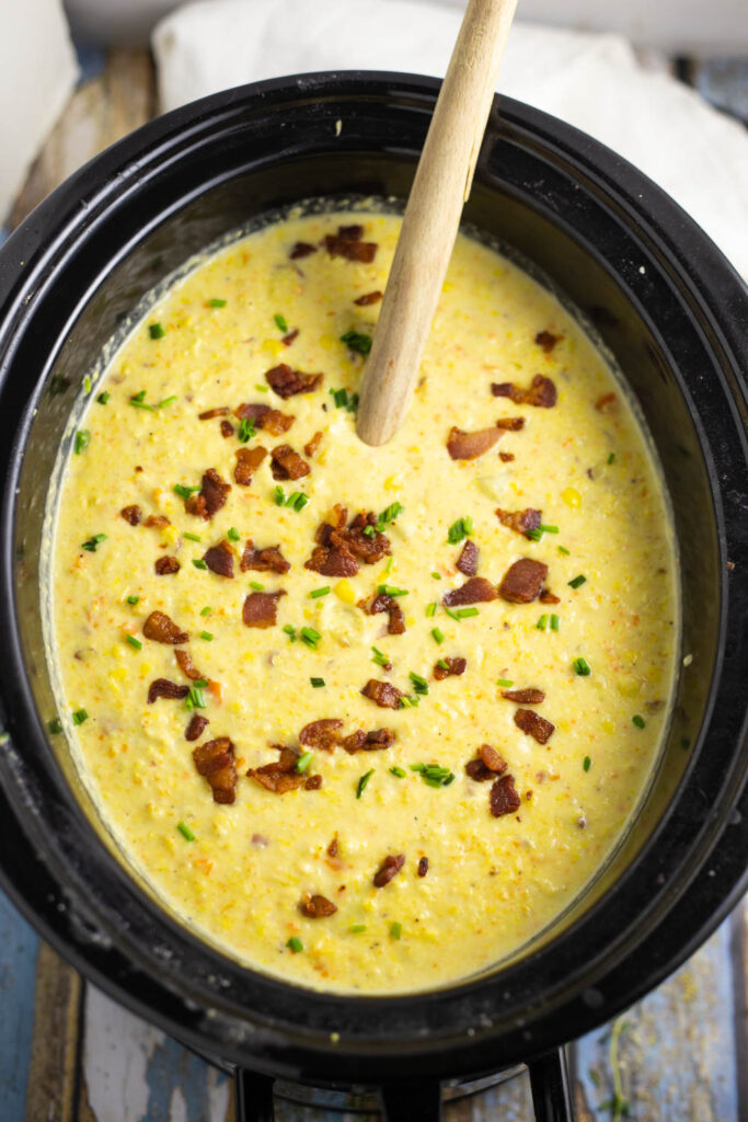 Crock Pot Corn Chowder in a slow cooker on a rustic wood background with a wooden spoon it, topped with crumbled bacon and chives