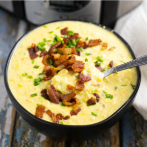 Black ceramic bowl filled with Crock Pot Corn Chowder with a spoon in it, topped with crumbled bacon and chopped chives.