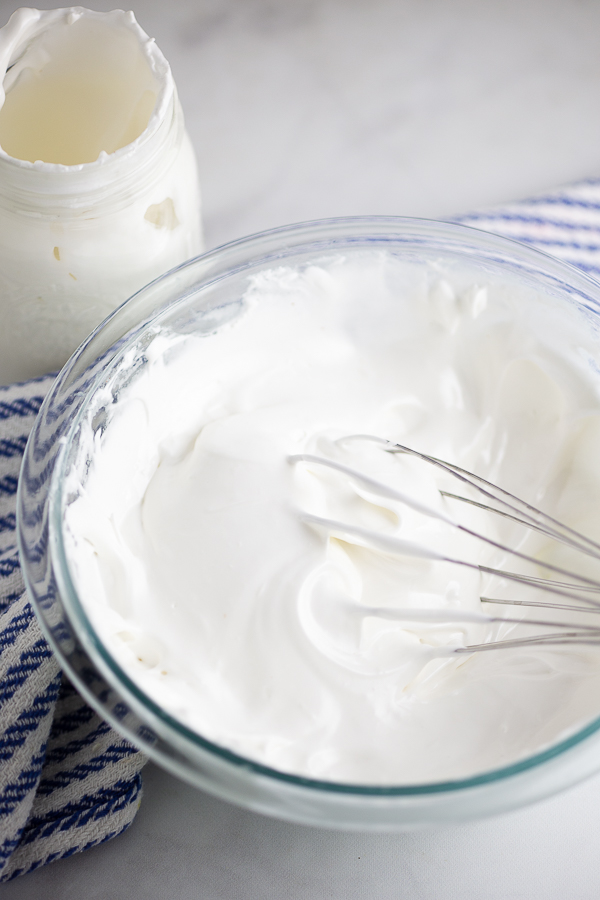 Homemade marshmallow fluff in a glass bowl with a whisk in it and a mason jar behind it.