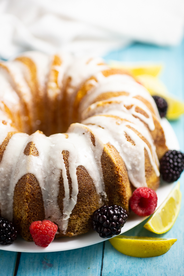 Side view of a Lemon buttermilk pound cake with lemon glaze with a linen behind it. The cake is surrounded by fresh raspberries and blackberries and lemon wedges.