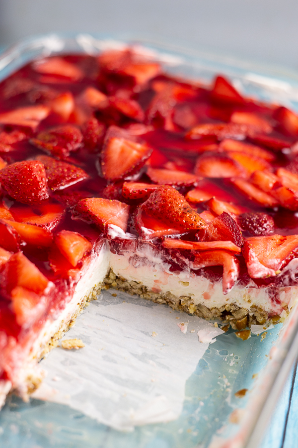 Strawberry pretzel salad in a glass casserole dish with several pieces cut out so you can see the crust, cream cheese, and strawberry layers.
