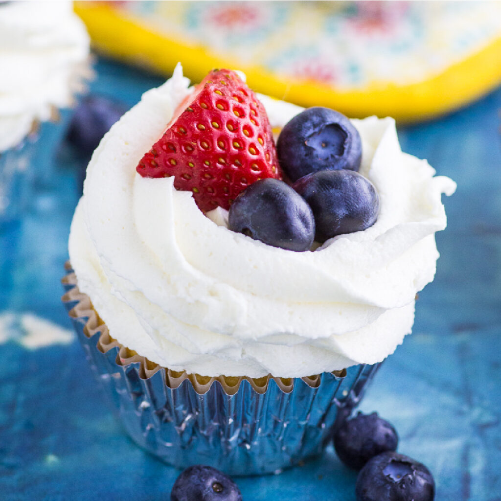 Berries and cream cupcake in a foil wrapper on a distressed blue cement backdrop, topped with whipped cream cheese frosting, half a fresh strawberry, and 3 fresh blueberries