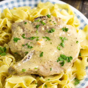 Two Crock Pot Ranch Pork chops garnished with fresh parsley on a bed of buttery egg noodles in front of a slow cooker.