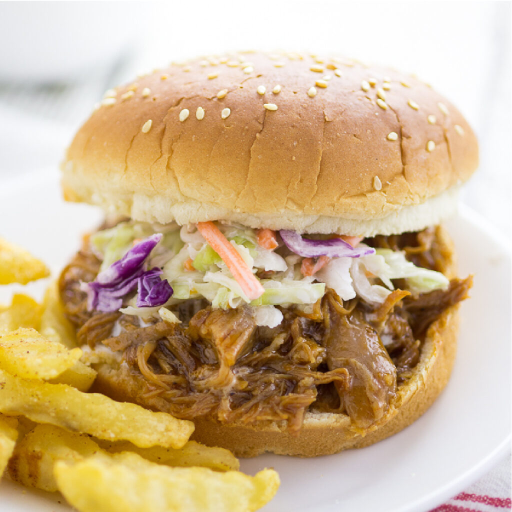 Easy Slow Cooker BBQ Pulled Pork topped with homemade coleslaw on a sesame seed bun next to crinkle cut fries on a small plate
