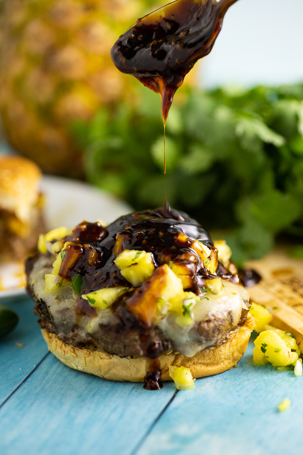 Teriyaki sauce being drizzled onto a burger topped with pineapple salsa.