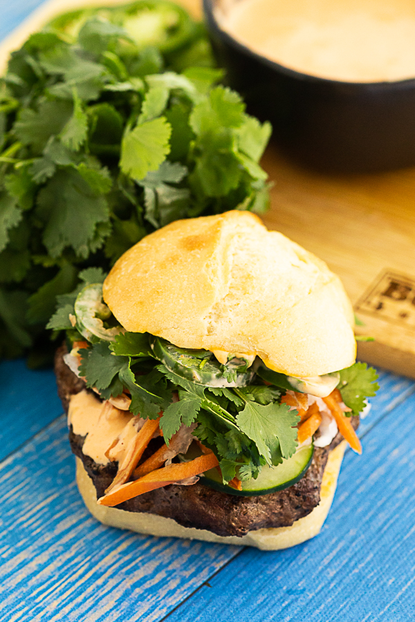 An overhead view of a fully topped banh mi burger in front of a cutting board, a bunch of fresh cilantro, and a bowl of spicy mayo.