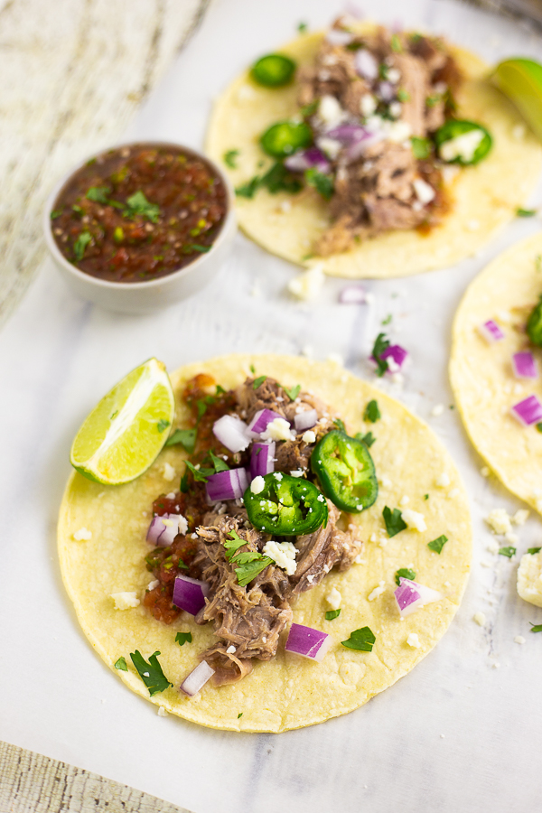 Carnitas on a corn tortilla topped with fresh jalapeno slices, red onion, cilantro, cotija cheese, and a lime wedge with a cup of salsa and 2 more tacos behind it.