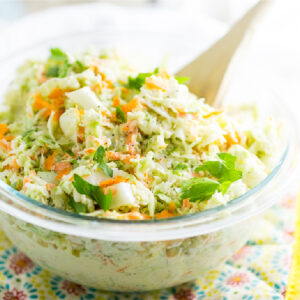 Creamy coleslaw in a clear glass bowl on a yellow flower pot holder with a wooden spoon sticking out