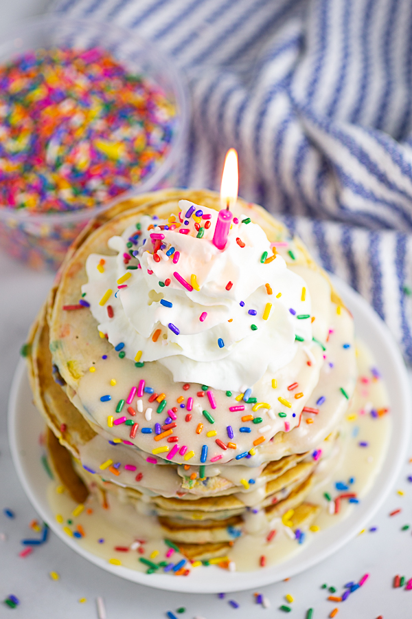 Overhead view of a stack of funfetti pancakes topped with vanilla glaze, whipped cream, rainbow sprinkles, and a candle, with more rainbow sprinkles in the background.