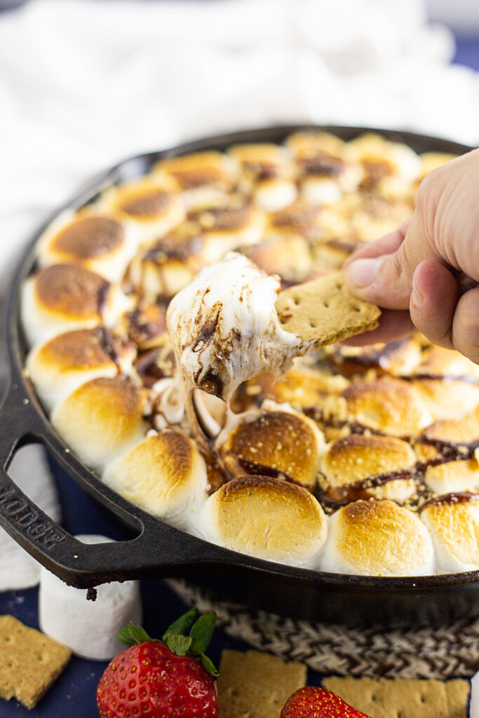 A graham cracker dipping into a cast iron skillet of gooey s'mores dip with a white linen napkin in the background.