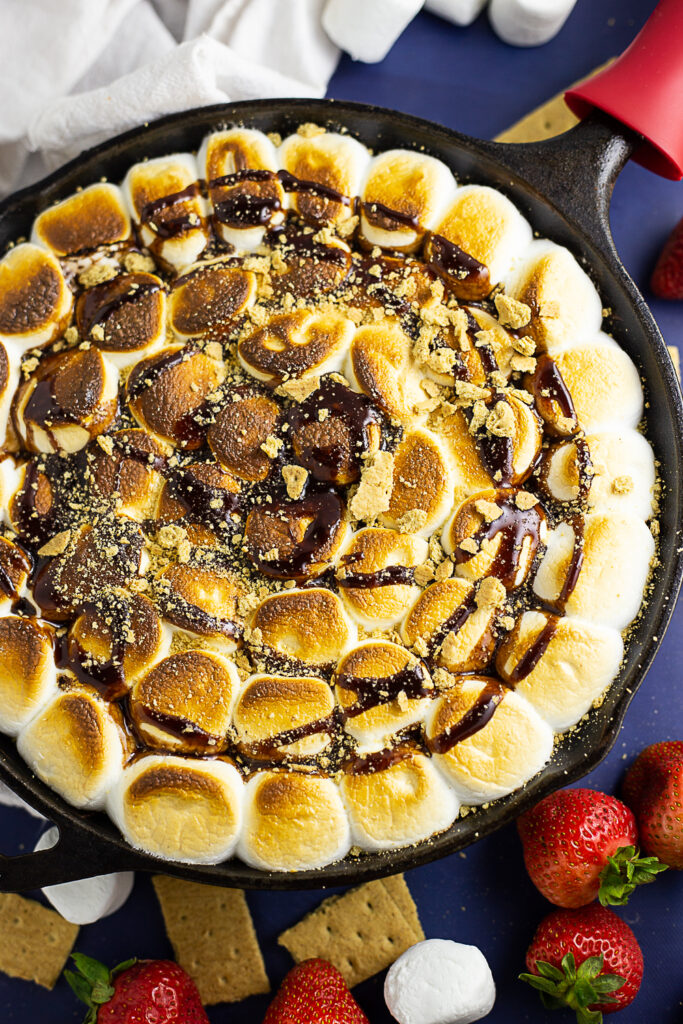 Overhead view of s'mores dip in a cast iron skillet next to a linen napkin and surrounded by graham crackers, marshmallows, and fresh strawberries.