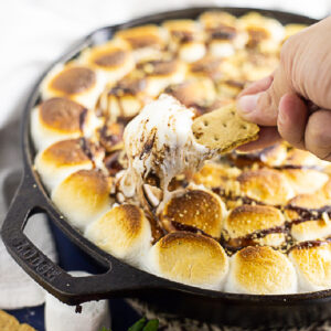 A graham cracker dipping into a cast iron skillet of gooey s'mores dip.