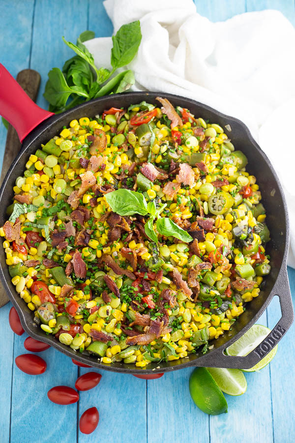 Overhead view of southern succotash in a cast iron skillet topped with bacon and fresh basil surrounded by a clean linen, lime wedges, more fresh basil, and cherry tomatoes