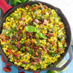 Overhead view of southern succotash in a cast iron skillet topped with bacon and fresh basil surrounded by a clean linen, lime wedges, more fresh basil, and cherry tomatoes