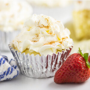 Tres Leches Cupcake topped with a sprinkle of cinnamon in a foil wrapper next to a fresh strawberry
