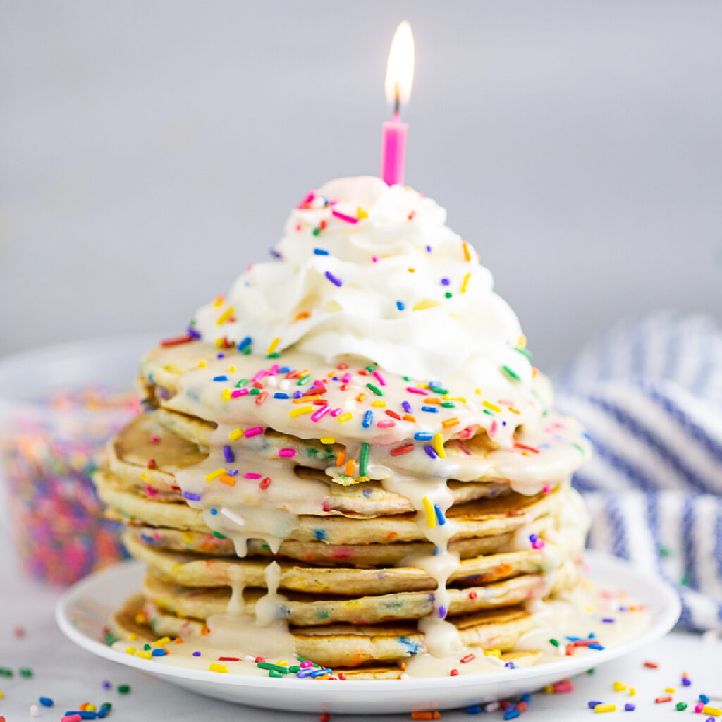 Funfetti pancakes drizzled with a vanilla glaze, topped with whipped cream, rainbow sprinkles, and a candle