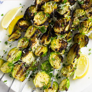 Grilled brussels sprouts on skewers on a plate with lemon wedges topped with grated Parmesan