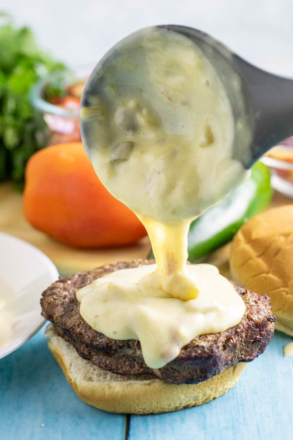 Queso being drizzled on top of a burger with tomato and jalapeno behind it.