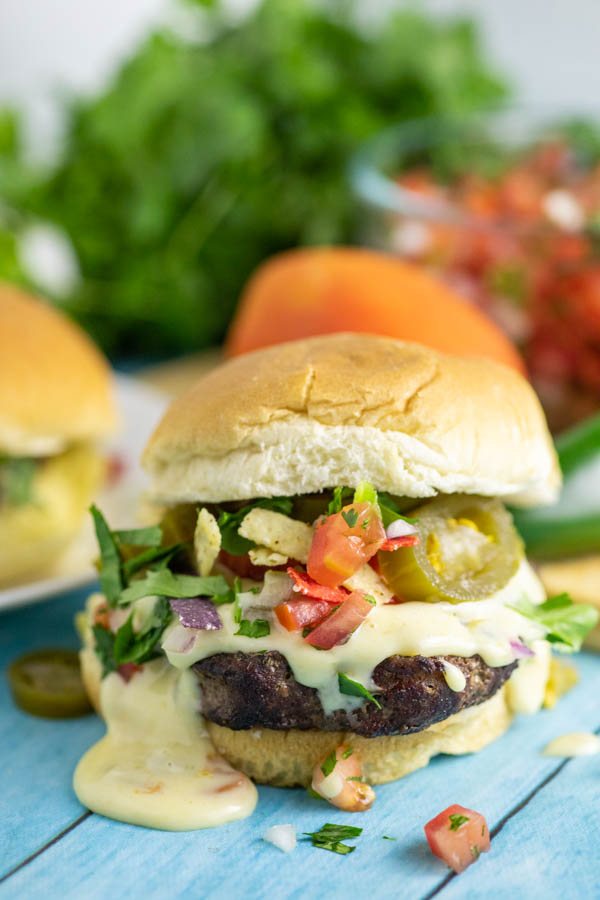 Close up of a nacho burger topped with queso, tortilla strips and jalapenos with fresh cilantro and another burger behind it.
