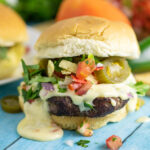 Close up of a nacho burger topped with queso, tortilla strips and jalapenos with fresh cilantro and another burger behind it.