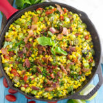 Overhead view of southern succotash in a cast iron skillet topped with bacon and fresh basil surrounded by a clean linen, lime wedges, more fresh basil, and cherry tomatoes
