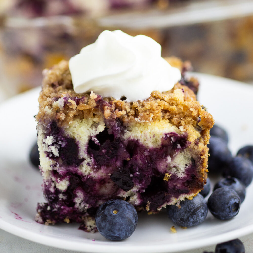 side view of blueberry buckle topped with whipped cream on a small plate with fresh blueberries