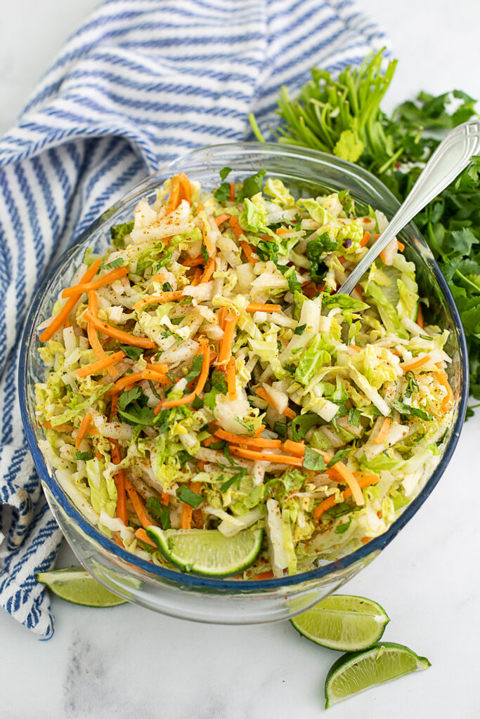 Overhead view of jicama slaw in a glass bowl with a spoon in it, garnished with lime wedges and next to a striped linen and fresh cilantro.