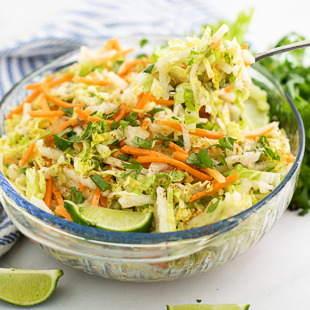 Large glass bowl filled with jicama slaw with a metal spoon taking a scoop out of it, surrounded by a lime wedge, a striped linen, and fresh cilantro.