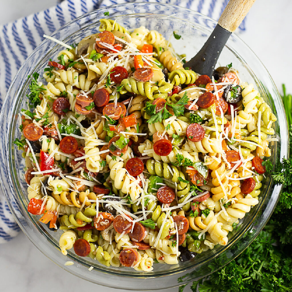 Overhead view of a large bowl of pizza pasta salad with a wooden spoon in it and a striped linen and a bunch of fresh parsley in the background.