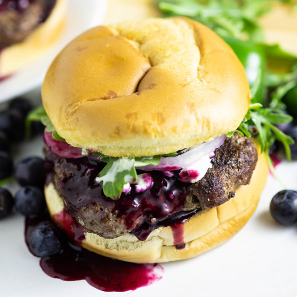 Spicy blueberry burger with blueberry sauce running down with fresh arugula and blueberries behind it
