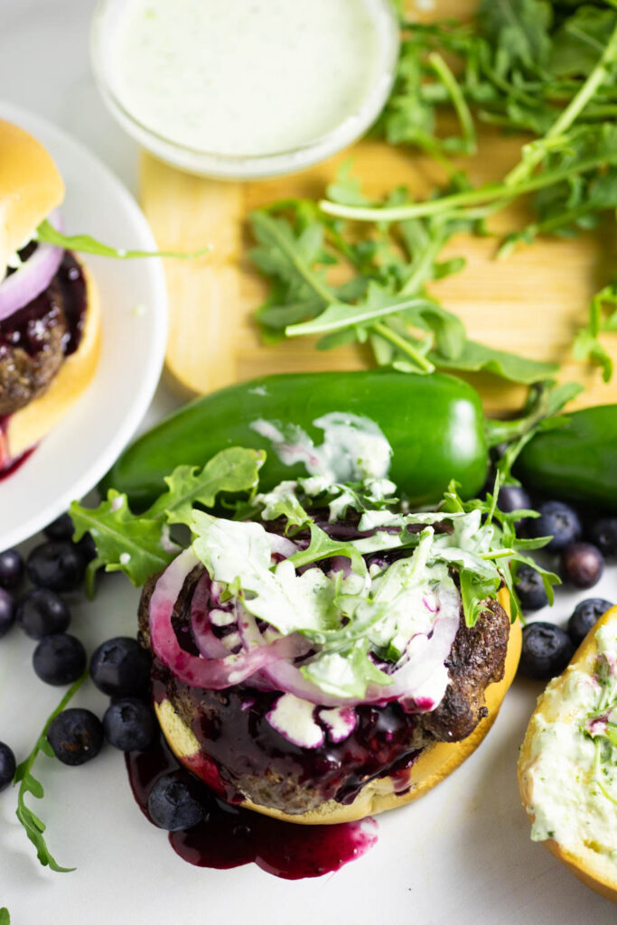 Open face spicy blueberry burger with blueberry sauce dripping down topped with jalapeno aioli, red onion, feta, and arugula surrounded by arugula on a wooden cutting board and fresh jalapenos and blueberries.