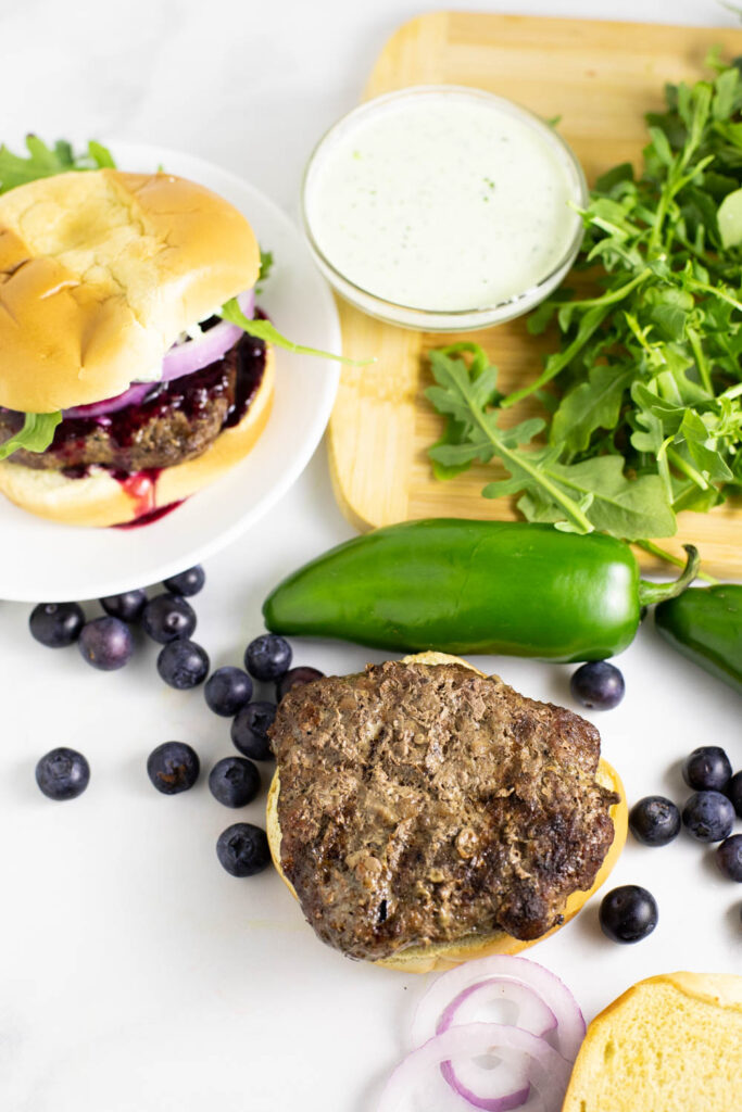 Ingredients to assemble spicy blueberry burgers including a burger on a bottom bun, red onion slices, blueberries, jalapeno, prepared jalapeno aioli, and arugula on a wooden cutting board