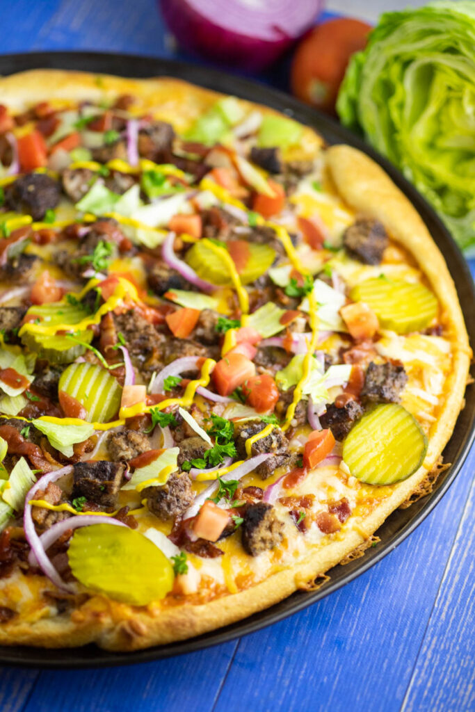 Close up side view of a bacon cheeseburger pizza on a blue wooden background with a wedge of iceberg lettuce, fresh tomato, and red onion in the background