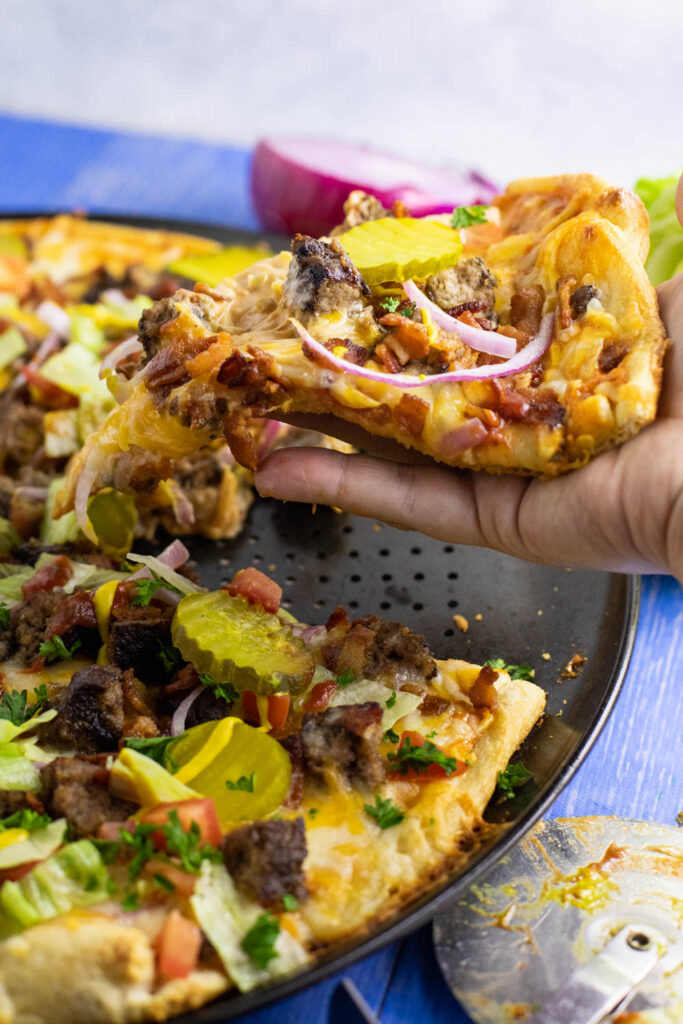 Hand pulling a slice of bacon cheeseburger pizza from the full pizza