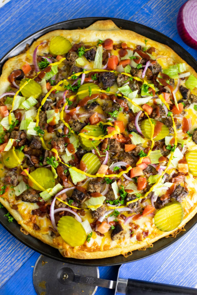 Overhead view of a bacon cheeseburger pizza next to a pizza cutter and red onion on a blue wooden background.