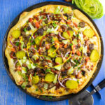 Overhead view of a full bacon cheeseburger pizza next to a pizza cutter and half a head of iceberg lettuce on a blue wood background