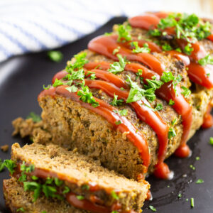 Full meatloaf on a large plate with 2 slices cut off garnished with ketchup drizzle and fresh chopped parsley.