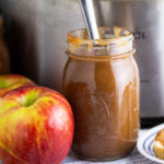 A jar of apple butter next to honeycrisp apples in front of a stainless steel Crock Pot.