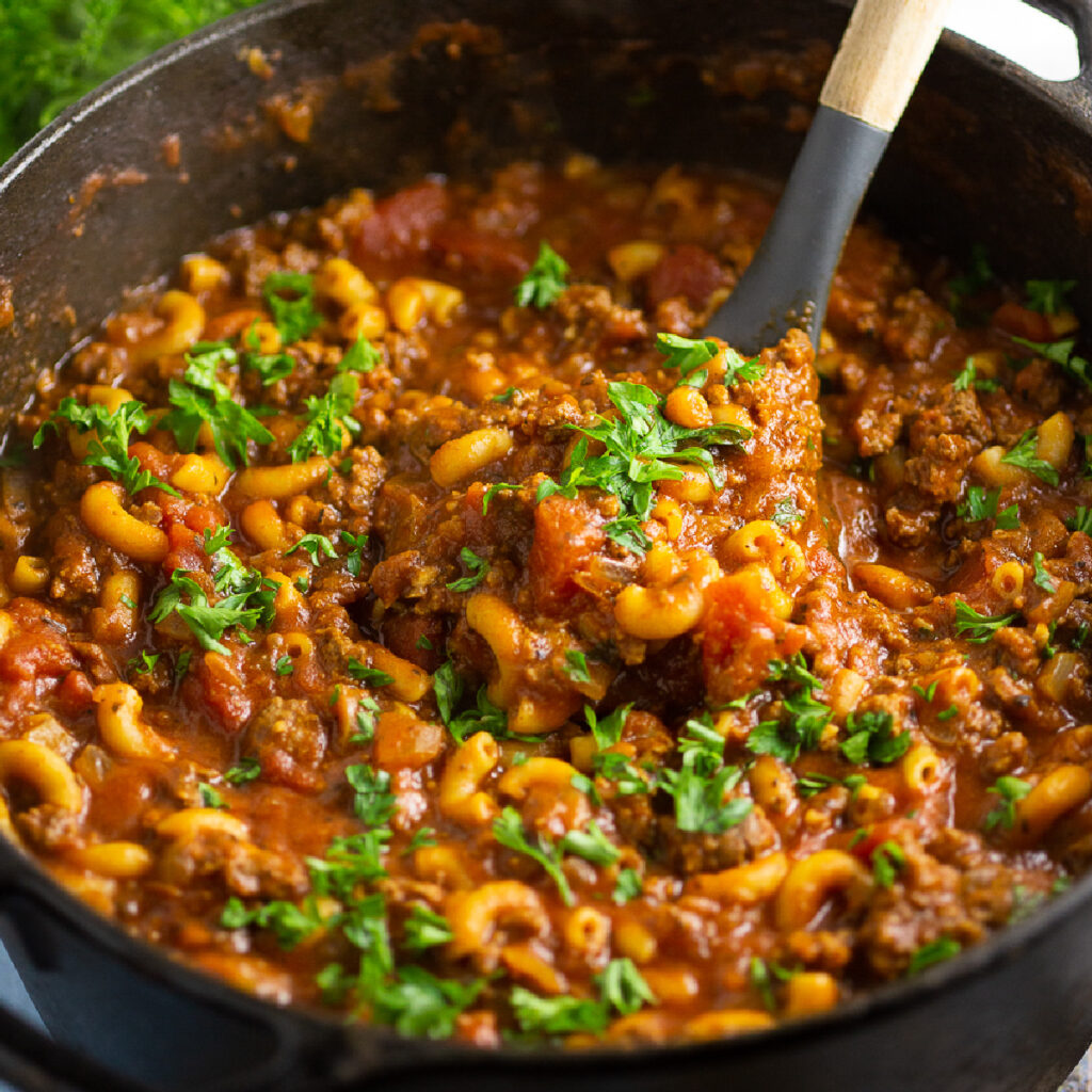 American goulash topped with fresh chopped parsley in a large cast iron Dutch oven with a wooden spoon scooping in the center.