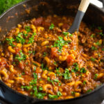 American goulash topped with fresh chopped parsley in a large cast iron Dutch oven with a wooden spoon scooping in the center.