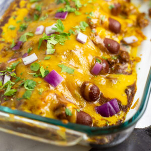 Chili cheese dog casserole in a glass 9-inch by 13-inch casserole dish topped with cilantro and red onions.