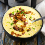 Black ceramic bowl filled with Crock Pot Corn Chowder with a spoon in it, topped with crumbled bacon and chopped chives.