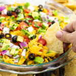 Easy layered taco dip in a round glass dish with a tortilla chip dipping into it