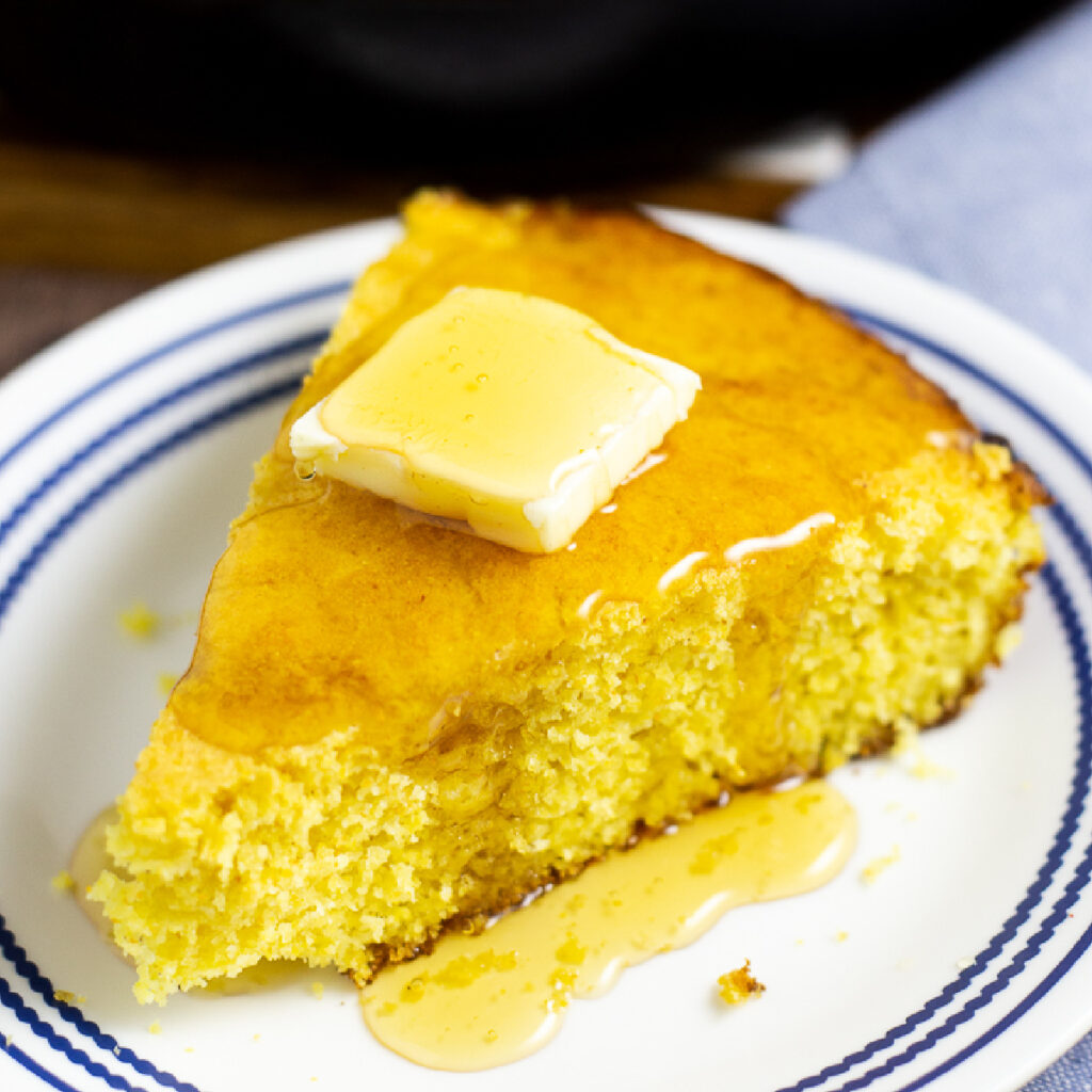 Wedge of cornbread with butter and honey on a white and blue plate on a rustic wood background.
