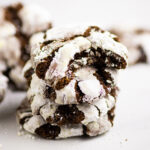 Stack of 4 chocolate crinkle cookies with a bite taken out of the top cookie on a plain white background with more cookies behind