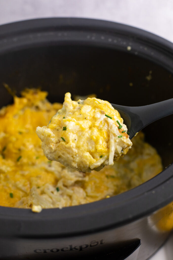 Large crock pot full of hash brown casserole with a silicone spoon scooping out a big portion.