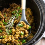 Crockpot Stuffing in a slow cooker with a wooden spoon in the middle topped with fresh parsley and rosemary.